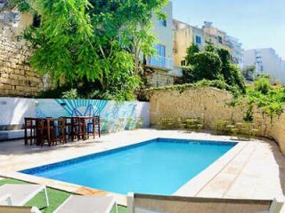 A small outdoor pool surrounded by a stone wall, greenery, and seating area with tables and chairs. Buildings are visible in the background.