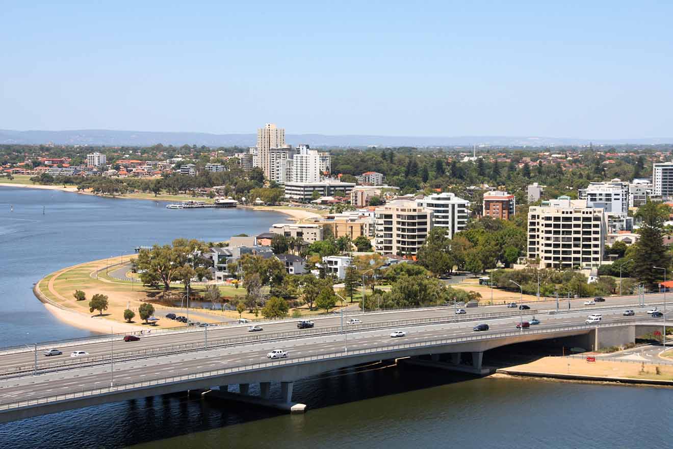 A bridge over a calm river leading to a cityscape, with clear skies and a mix of residential and commercial buildings