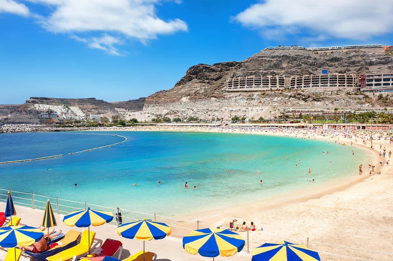 A beach with yellow and blue umbrellas, a curved bay with turquoise water, and numerous people sunbathing and swimming. Hills and buildings are in the background under a partly cloudy sky.