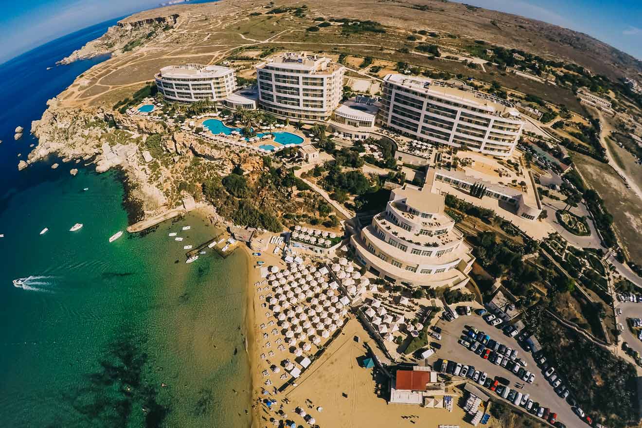 Aerial view of a seaside resort with multiple buildings, swimming pools, a sandy beach with umbrellas, and parked cars along the coastline.