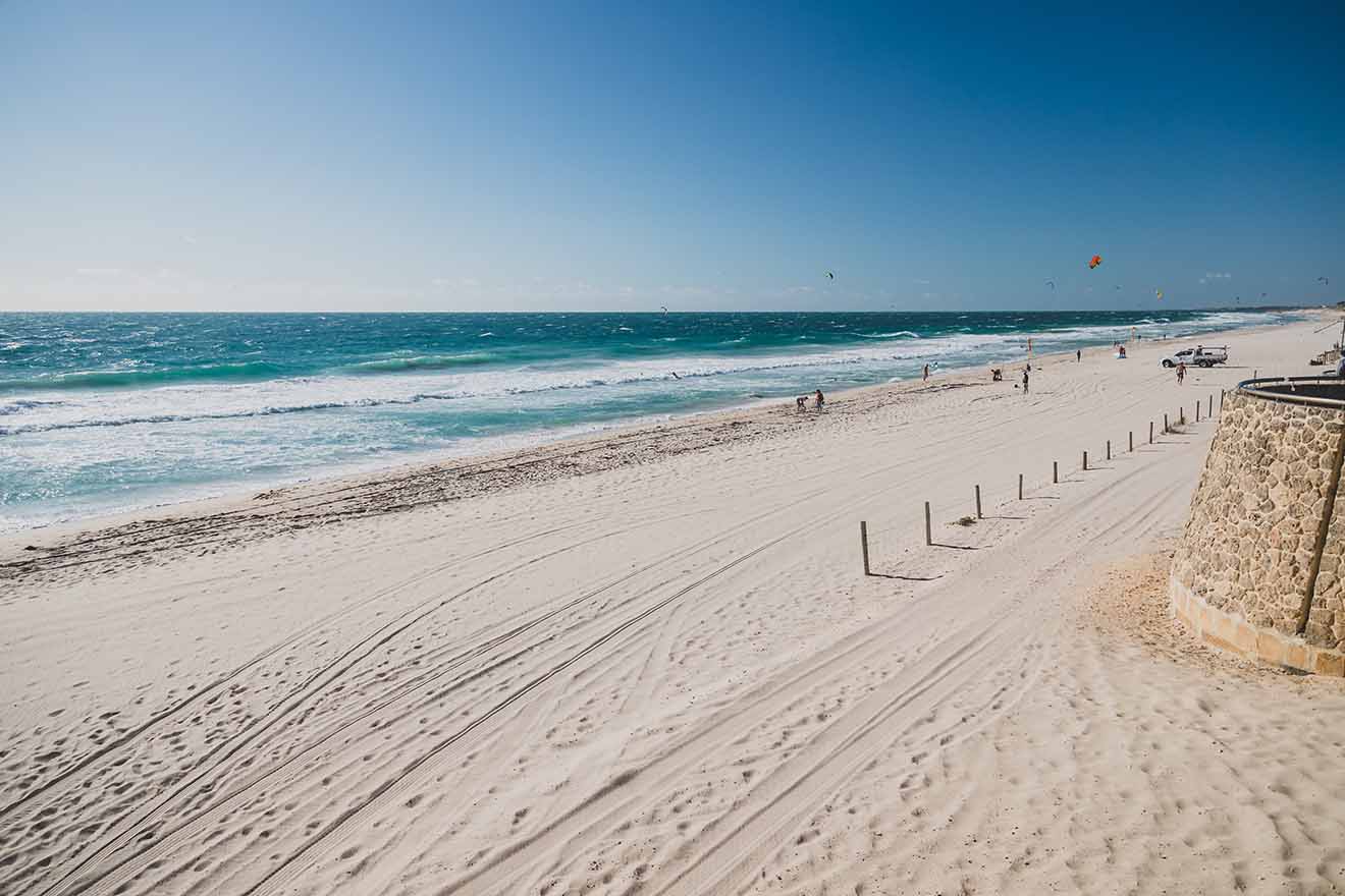 A wide sandy beach with a clear blue sky, kite surfers in the distance, and a calm sea