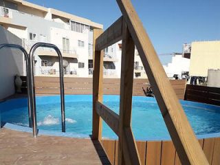 A rooftop terrace features a small round swimming pool with metal ladder and wooden railing, surrounded by residential buildings under a clear blue sky.