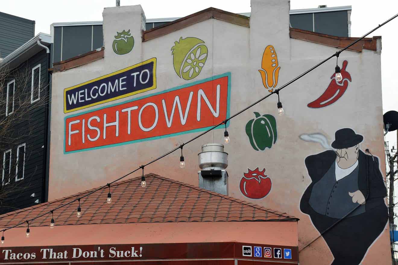 Colorful mural reading "Welcome to Fishtown" featuring vibrant images of a lemon, corn, and other motifs, with "Tacos That Don't Suck!" slogan below