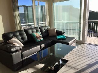 A black leather sectional sofa with cushions is in a sunlit room, facing a glass coffee table. Large windows offer a view of the mountains and a balcony with white railings.