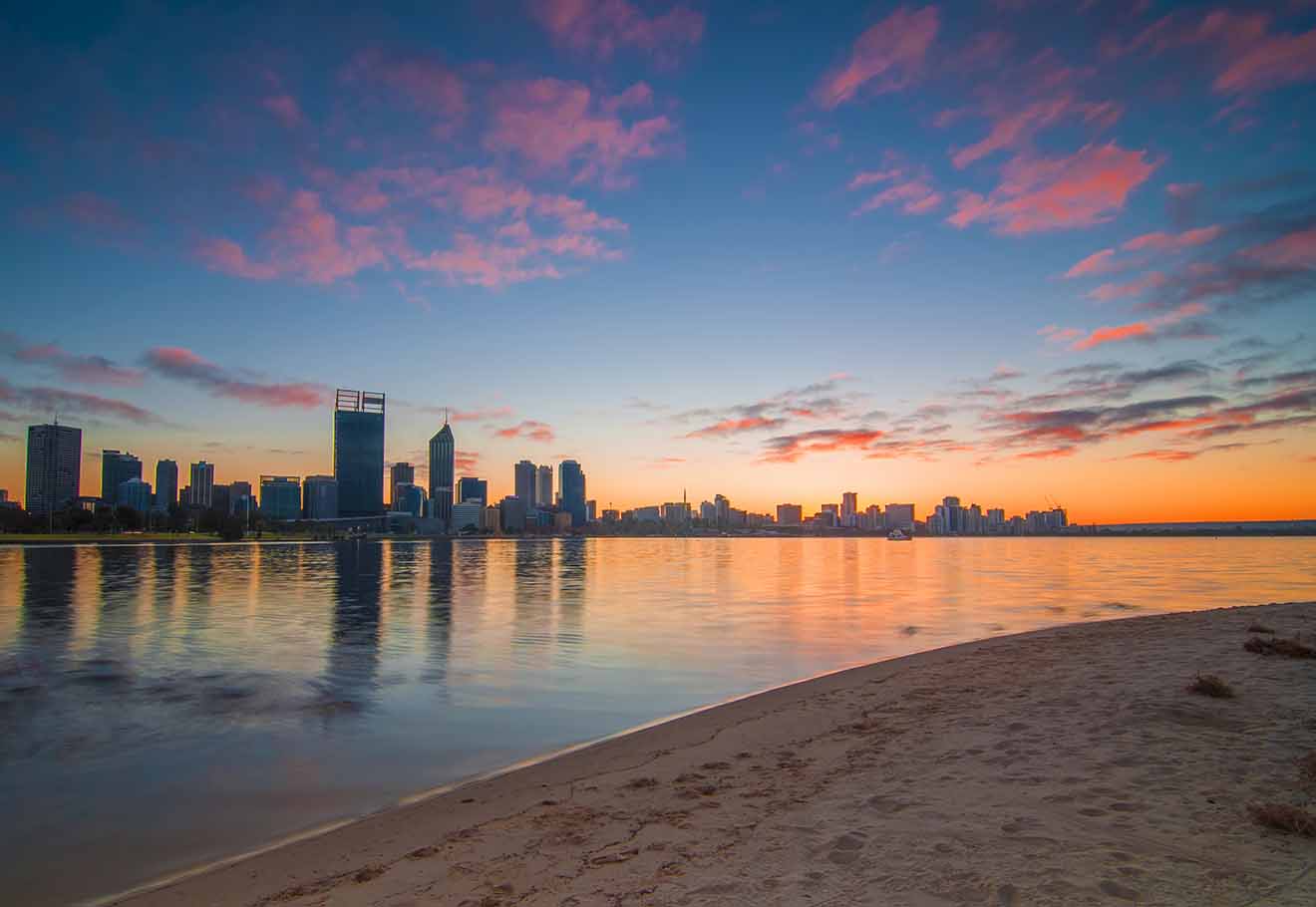 A sunset city beach scene with a glowing sky reflecting on water and silhouettes of city buildings in the backdrop