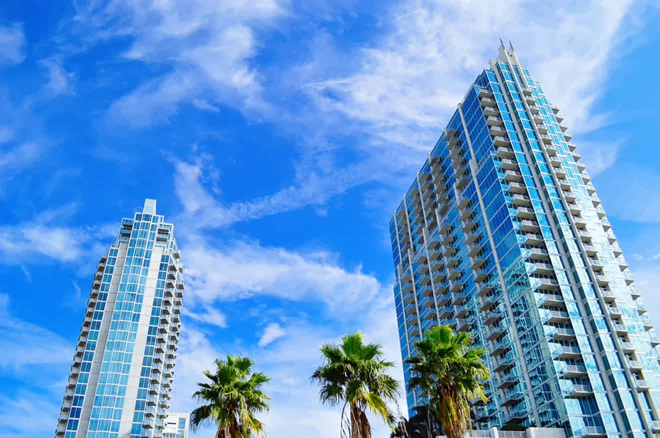 Two modern high-rise condominiums towering over palm trees against a wispy clouded blue sky