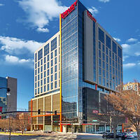 A modern multi-story hotel building with large glass windows, located on a city street corner under a blue sky with scattered clouds.