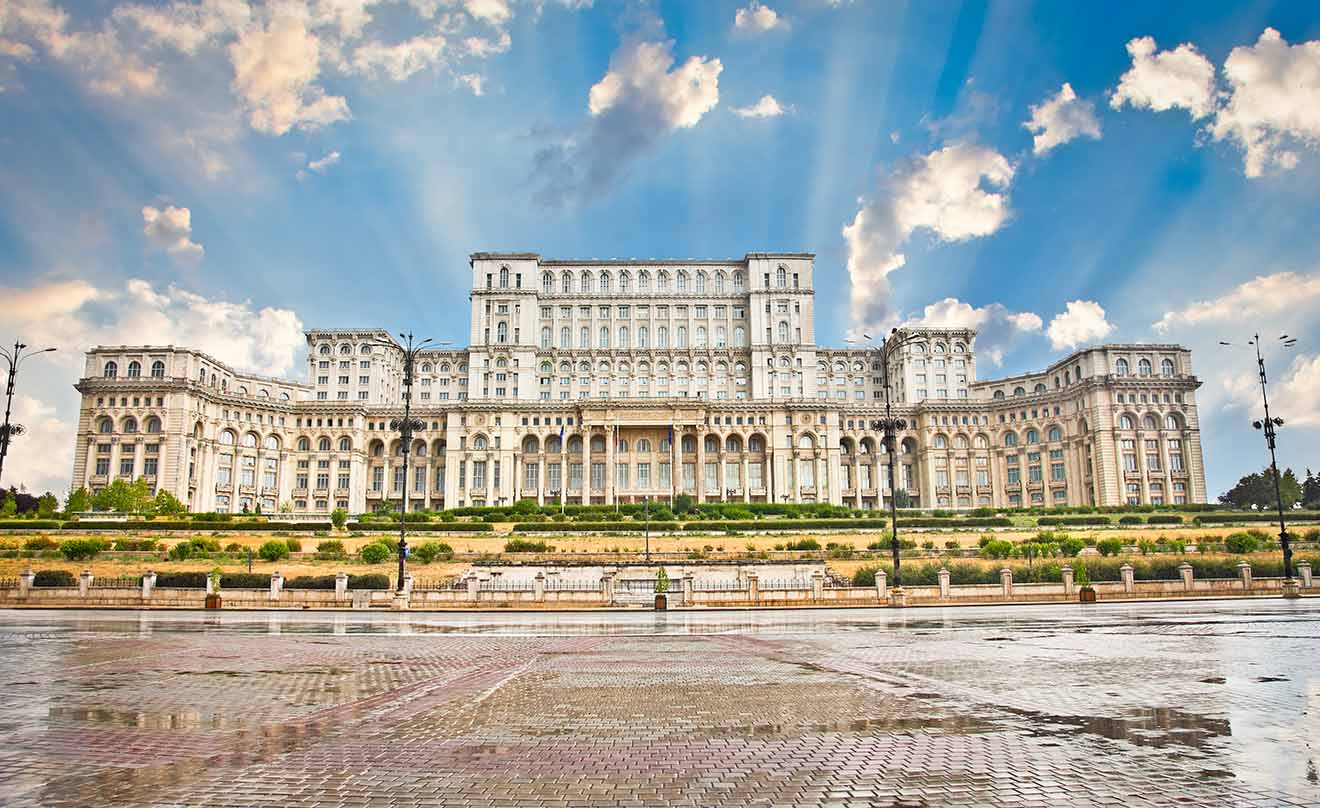 The Palace of the Parliament in Bucharest, Romania, under a dramatic sky with sunbeams shining through the clouds, highlighting the grandeur of this massive, imposing government building