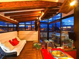 A cozy rooftop apartment interior at dusk with wooden beams, a sofa bed, a kitchenette with a red countertop, and a terrace overlooking the cityscape