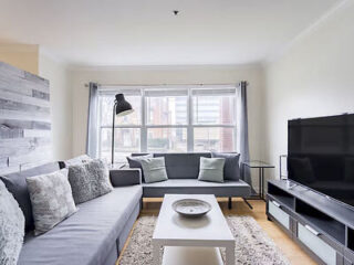 Modern living room with gray sofas, a white coffee table, a floor lamp, a large flat-screen TV, and a window with sheer curtains letting in natural light.