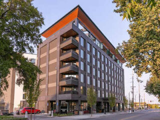 A modern six-story apartment building with balconies, a distinctive angled roof, and street-level commercial space, surrounded by trees and parked cars.