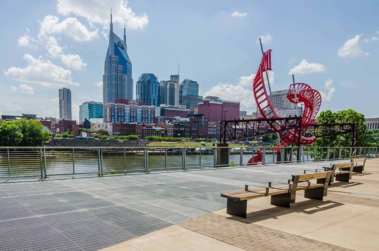 Nashville riverside featuring an artistic red sculpture with the city skyline and the AT&T Building, also known as the Batman Building, in the background