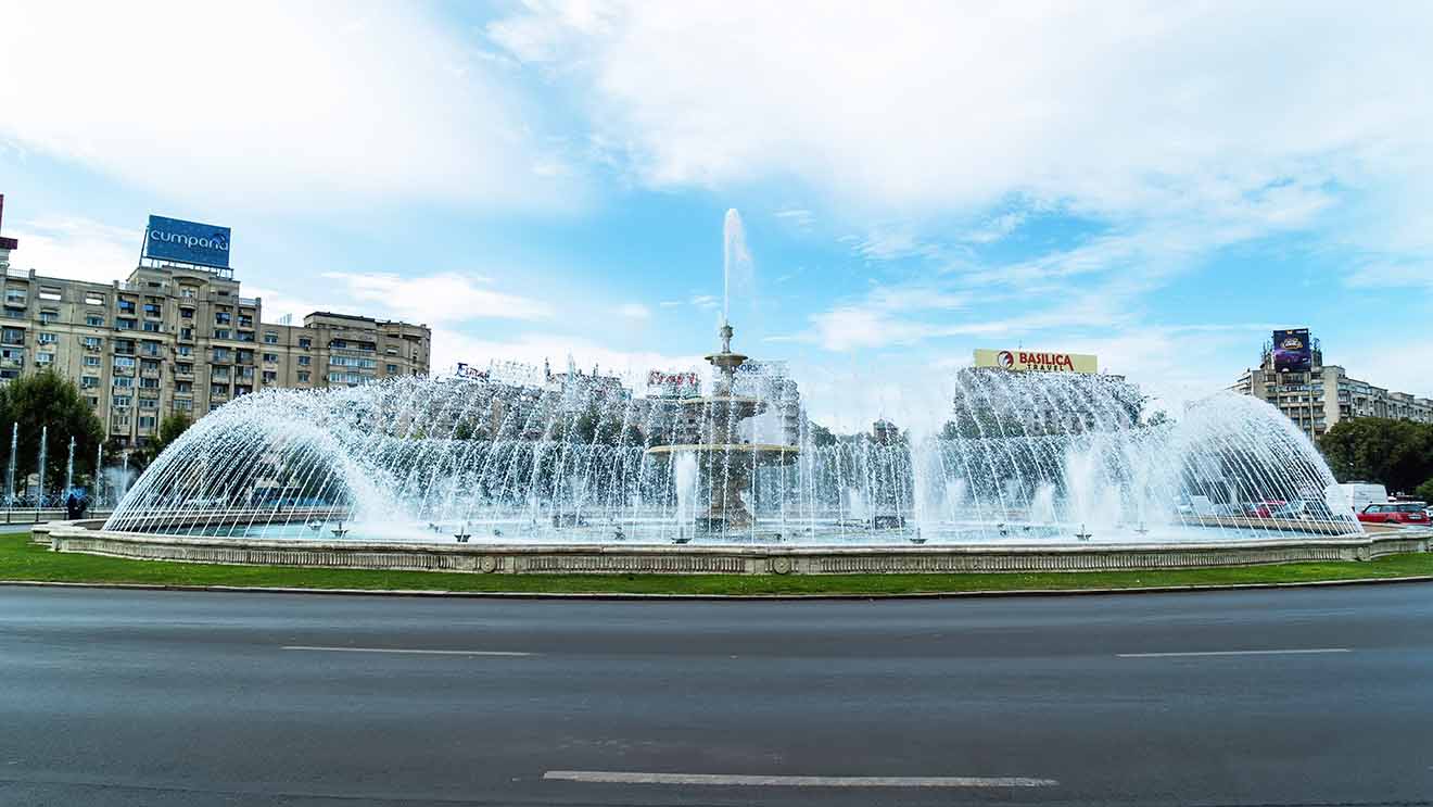 A large, ornate fountain located in a busy city roundabout, featuring multiple jets of water creating a spectacular display against a backdrop of commercial buildings and clear blue skies