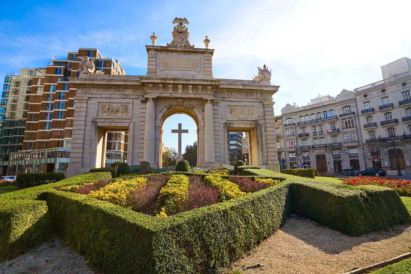 The grand Puerta de Alcalá, a neoclassical monument in Madrid, Spain, with intricate sculptures and reliefs, flanked by lush garden hedges and contrasting modern buildings, under a clear blue sky