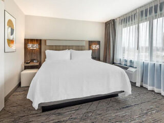 Modern hotel room with a large bed, white bedding, two bedside lamps, a large window with sheer curtains, and abstract wall art.
