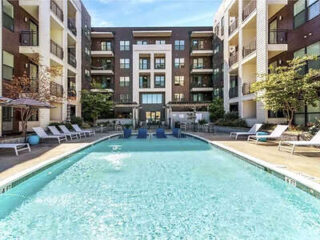 Rectangular outdoor swimming pool surrounded by lounge chairs and umbrellas, with a modern multi-story apartment building in the background.