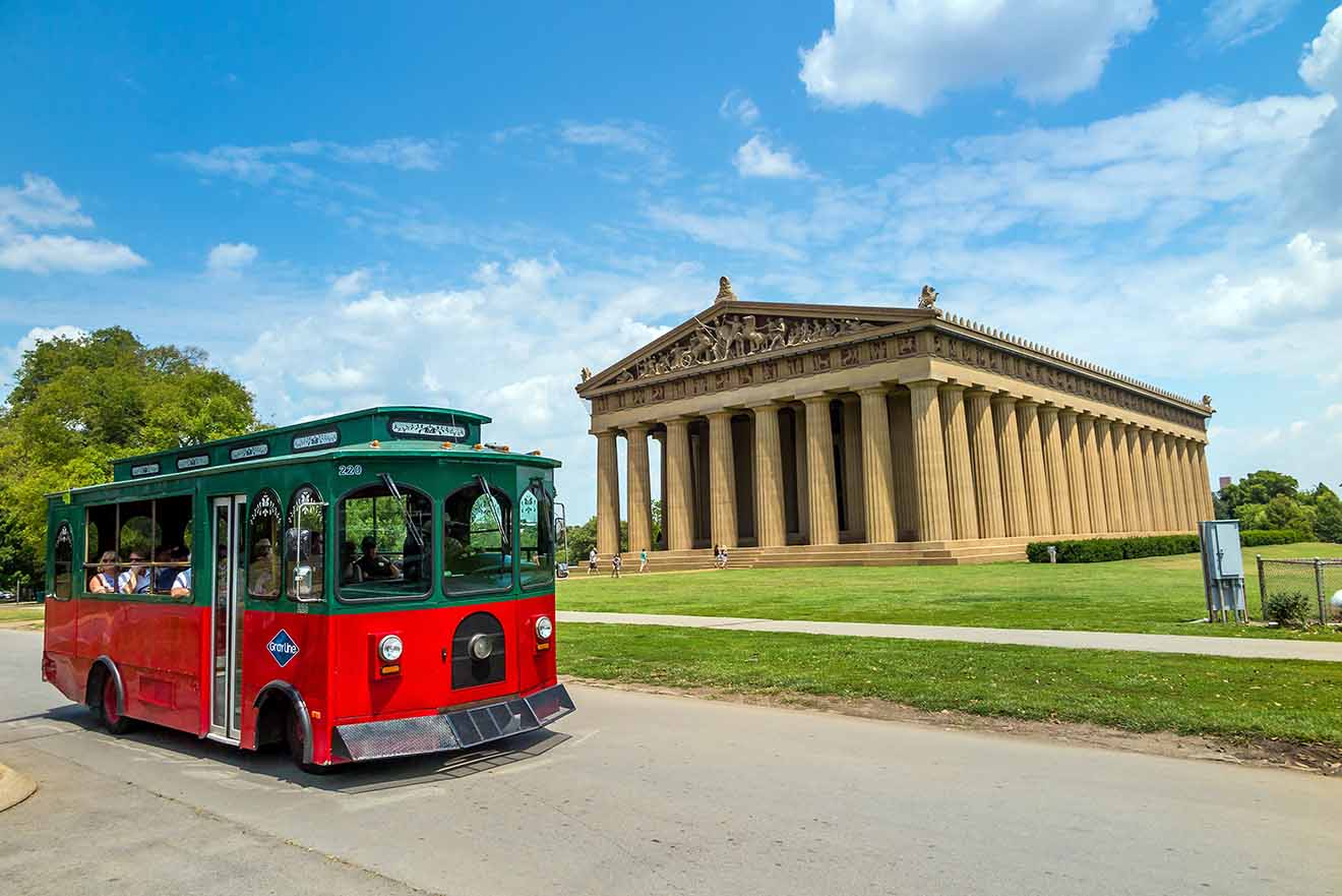 A red trolley tour bus passing in front of the full-scale replica of the Parthenon in Nashville's Centennial Park under a blue sky with fluffy clouds