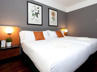 A well-appointed hotel room with a large bed with white linens and orange accent pillows, flanked by two nightstands with lamps, and wall art above the headboard