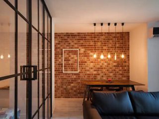 A chic urban apartment living room with a black sofa, a metal framed glass partition, exposed brick walls, and modern hanging light fixtures.