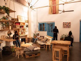 A casual and artistic co-working space with eclectic furnishings, including a wooden pallet sofa with cushions, bookshelves, and artwork