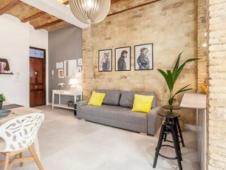 A modern and chic living room in an urban apartment with exposed brick walls, a gray sofa with yellow accent pillows, a wall-mounted TV, and a dining area with stylish chairs and artwork