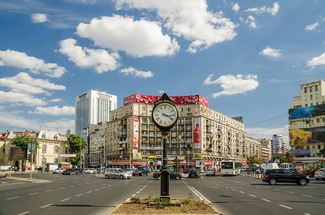 A busy urban intersection with a vintage street clock in the foreground, surrounded by traffic and commercial buildings under a partly cloudy sky