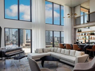 Modern living room with floor-to-ceiling windows, city views, a white sectional sofa, a black piano, and a stylish bar area with shelves and brown barstools.