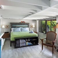 A bedroom with a large bed, green blanket, two bedside lamps, a chair, bench with shelves, and large windows showing greenery outside.