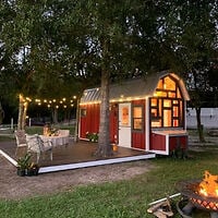 A small red tiny house with a deck, string lights, outdoor dining area, and a fire pit in front, surrounded by trees at dusk.