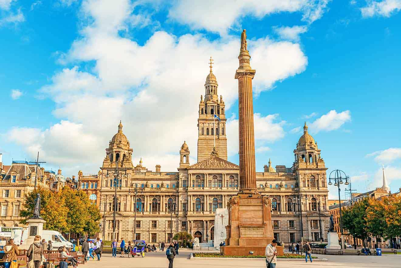 George Square in Glasgow on a sunny day, showcasing the grand Glasgow City Chambers and the prominent Sir Walter Scott monument, with people milling about in the square