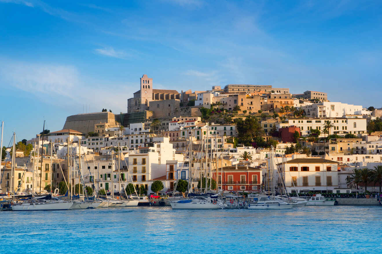 A coastal town with a marina filled with sailboats, white buildings, and a church perched on a hill under a clear blue sky.