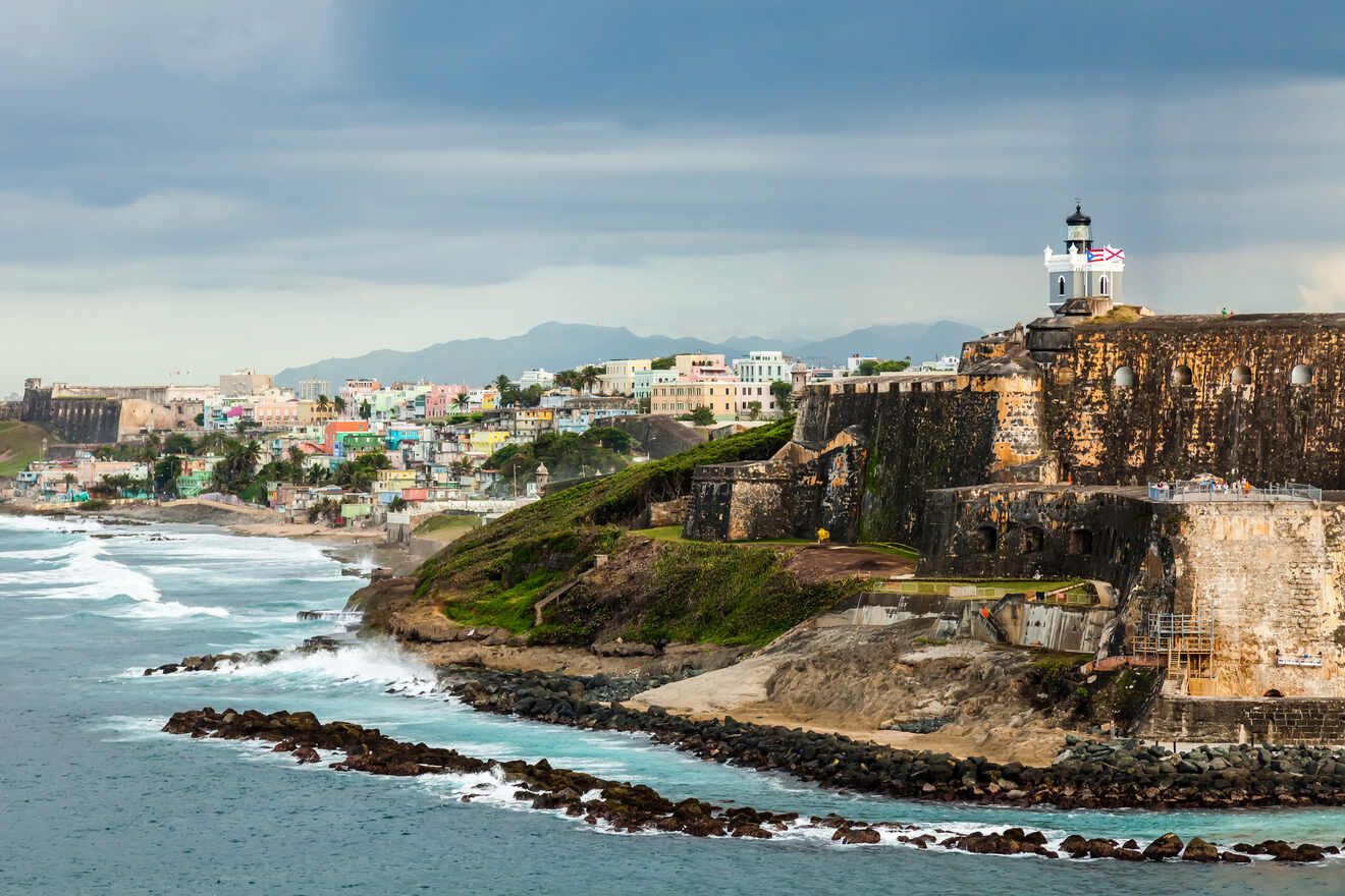 View of a coastal city with a historic fort, ocean waves, and colorful buildings under a cloudy sky.