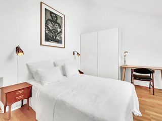 A minimalist bedroom with a single white bed, white bedding, a wooden nightstand, and a sleek, modern desk with a classic black chair under a wall-mounted lamp.