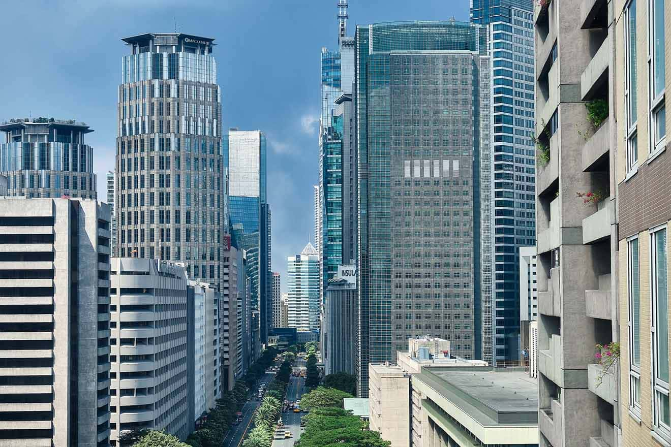 Street-level perspective of a tree-lined avenue in Makati, Manila, flanked by tall commercial buildings under a clear blue sky