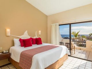 Hotel room with a large bed, red accents, and a balcony offering a view of a coastal town and the ocean.