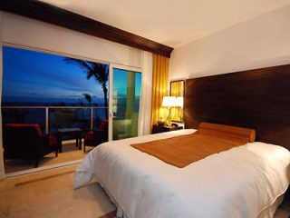 Luxurious bedroom with a wide window overlooking the ocean at sunset, featuring a minimalist design with dark wood tones and red accents.