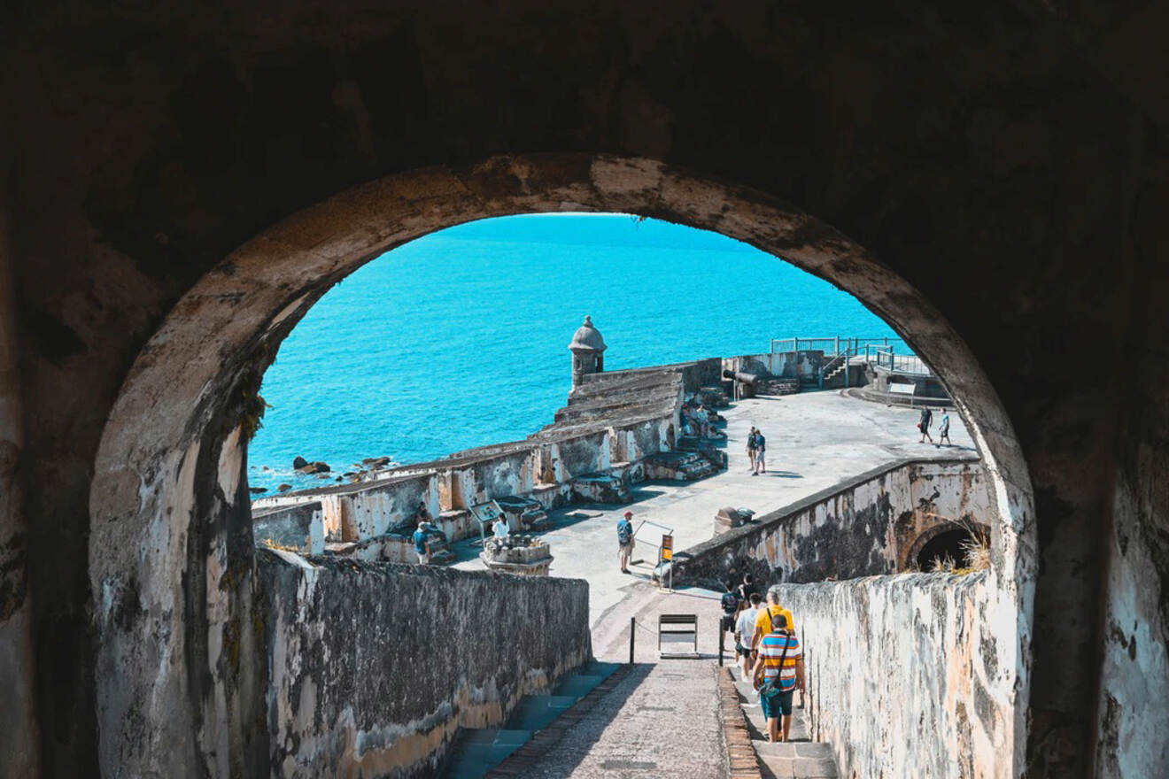 View through an archway overlooking a coastal fortification with people walking on a path, and the ocean in the background.