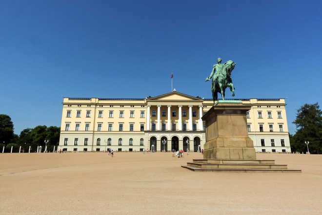 The Royal Palace in Oslo, Norway, on a sunny day with a clear blue sky, showcasing the grand yellow building and a prominent statue of King Karl Johan on horseback in the foreground.