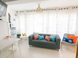 Modern and bright living space with a grey sofa, white dining set, and pops of color from cushions and decor, reflecting a clean, contemporary vacation home.