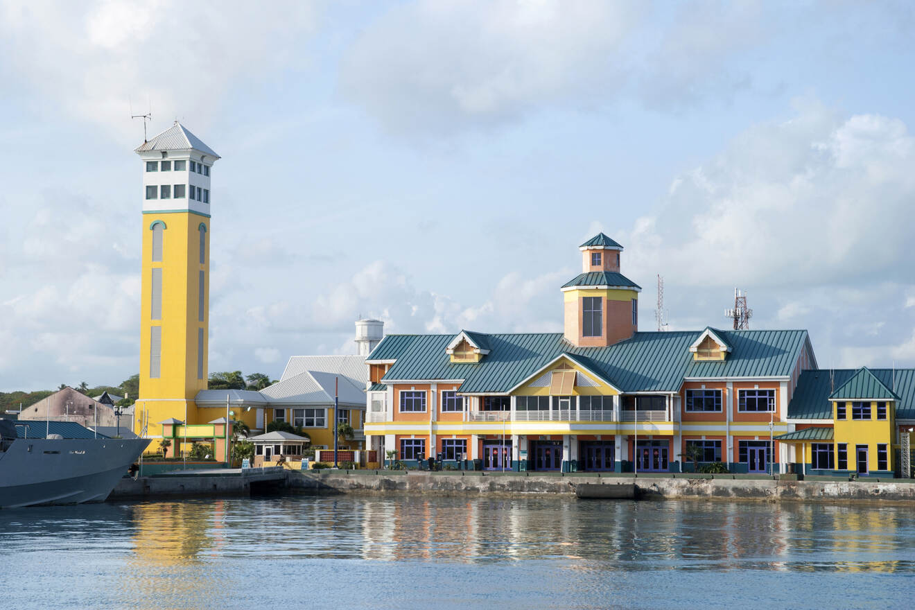 Waterfront view of a colorful marina in Nassau with a tall yellow tower, against a backdrop of cumulus clouds, reflecting a serene tropical setting.