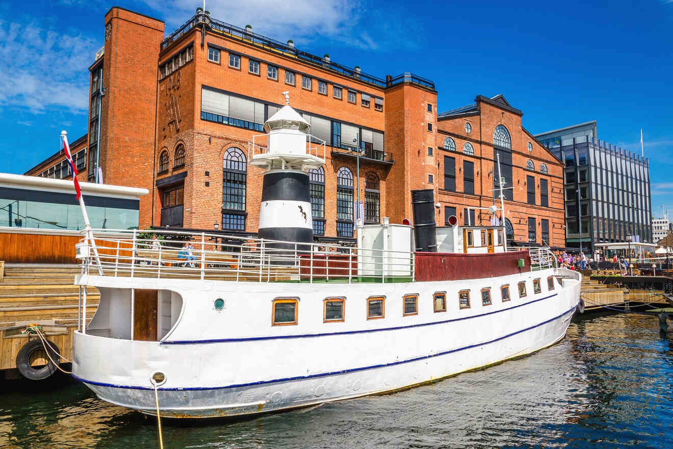 A vintage white ship docked at a harbor with brick buildings in the background, featuring a black and white lighthouse structure on its deck.