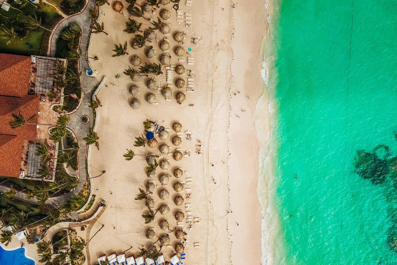 Aerial view of a resort beach with thatched umbrellas and a clear demarcation between the calm turquoise sea and the sandy shore.
