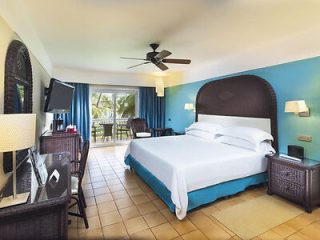 Spacious hotel room with a large bed, blue accents, and a balcony overlooking lush palm trees, epitomizing a luxurious tropical resort experience.