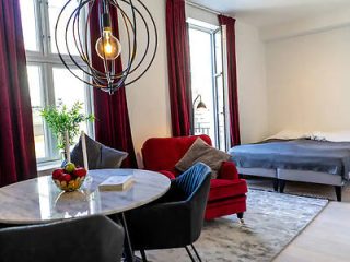 Elegant studio apartment with a grey round dining table, red velvet sofa, and a bed in the background, with a modern chandelier overhead.