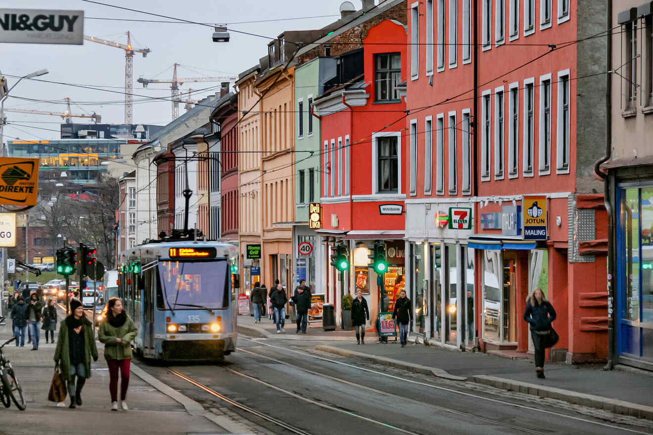 A busy street scene in Oslo, Norway, with pedestrians, a tram in motion, and colorful buildings lining the road under a cloudy sky.
