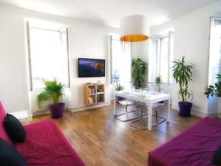 Bright and modern living room with hardwood floors, white walls, two purple couches, and a dining area with a white table, enhanced by natural light from large windows.