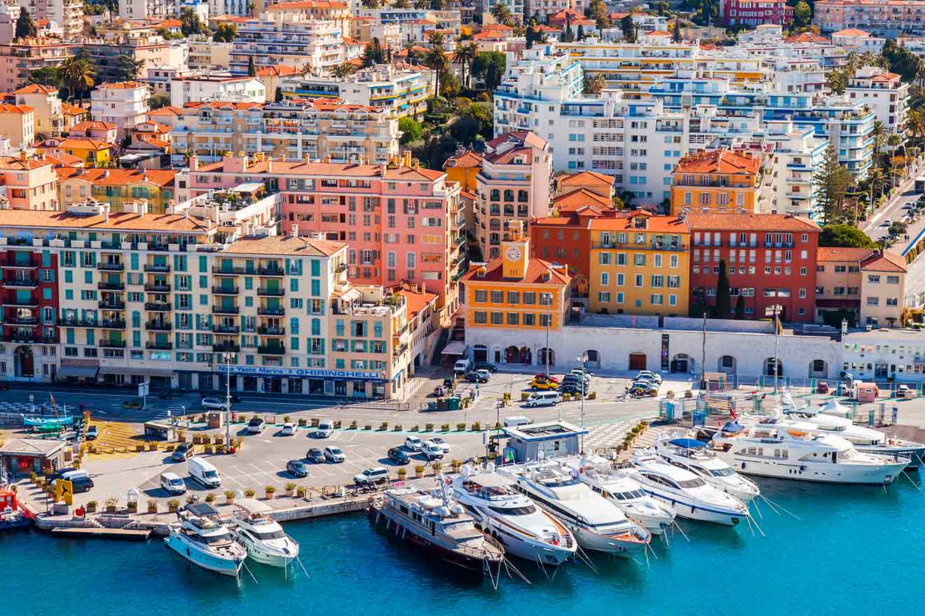 Panoramic view of a marina lined with multi-colored buildings, luxury yachts moored along the docks, and the bustling cityscape of Nice, France in the background