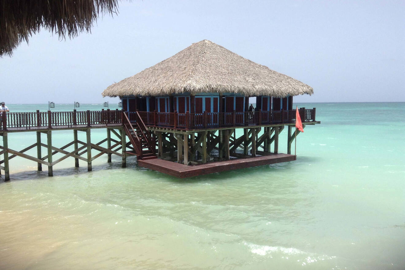 Overwater bungalow with a thatched roof extending over the serene turquoise sea, connected by a wooden jetty.
