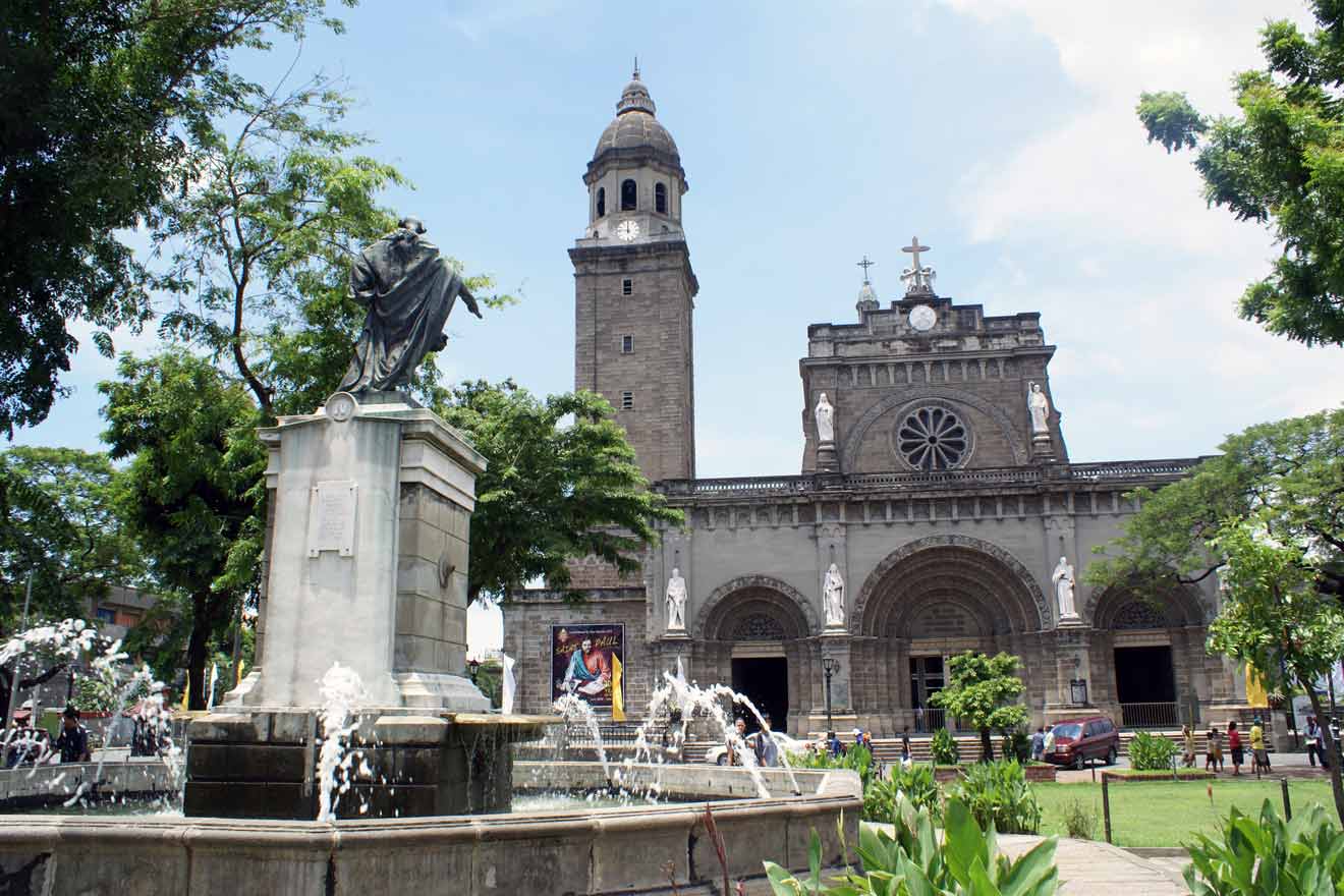 Historic Manila Cathedral with a prominent bell tower and a statue in the foreground, framed by lush trees and a clear sky