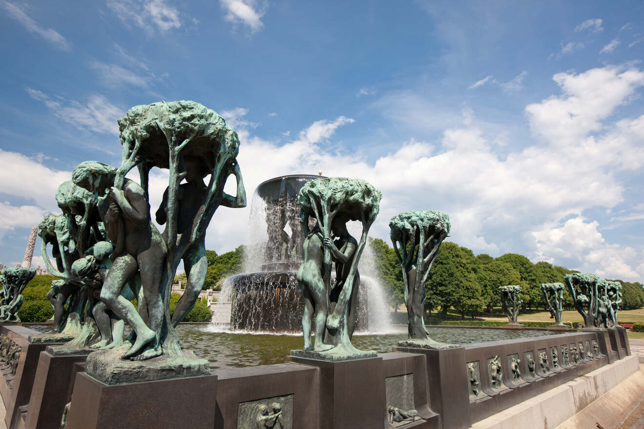 Sculptural fountain at Vigeland Park with multiple bronze figures beneath a tree canopy design against a bright blue sky with fluffy clouds.